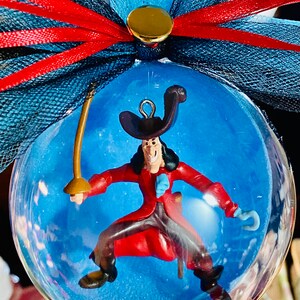 May include: A clear glass ornament with a blue background and a plastic figurine of Captain Hook inside. The ornament is decorated with a red and blue bow.
