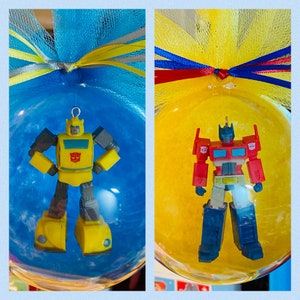 May include: Two Christmas ornaments featuring Transformers characters. The first ornament is yellow and features Bumblebee. The second ornament is red and features Optimus Prime.