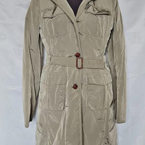 May include: A taupe-colored, knee-length coat with a smooth, slightly iridescent finish. It features a collared neckline, long sleeves, two chest pockets with flaps, two lower pockets with flaps, and a matching belt with a dark brown buckle.