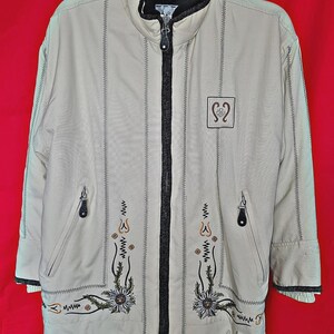 May include: A beige zip-up jacket featuring a black inner collar and trim. The front displays vertical stitched lines, two zippered pockets, and embroidered floral motifs with white flowers and green stems. A square patch with a symbol is on the chest.