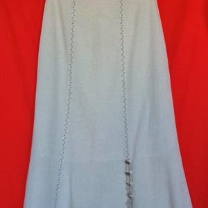 May include: A light beige, A-line skirt featuring vertical decorative stitching along the front panels. The skirt includes a side slit detail adorned with four small, round, metallic-toned buttons and a dangling cord accent at the hem.