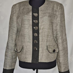 May include: A beige and grey textured jacket featuring a dark grey central placket with six silver-toned buttons. The jacket includes long sleeves with dark grey cuffs, a round neckline, and two front pockets with dark grey trim and button details.