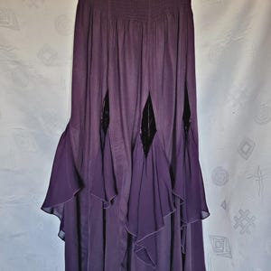 May include: A deep purple maxi skirt featuring a wide, smocked elastic waistband. The skirt has a layered design with vertical cutouts revealing dark, velvet-like fabric panels underneath, accented by cascading, ruffled fabric sections along the hem.
