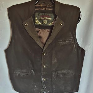 May include: A dark brown leather vest featuring a button-front closure with three buttons, two lower welt pockets, and one chest welt pocket. The interior label displays the text c.comberti. The vest has a stand-up collar and a brown lining.