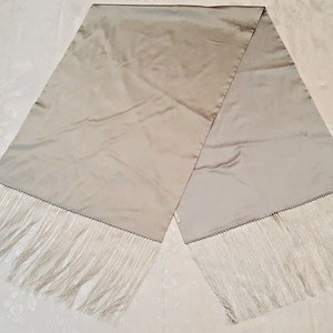 May include: A rectangular scarf in a solid light taupe or champagne color with a smooth, satin-like finish. The scarf features long, thin, matching light-colored fringe trim along both of the shorter ends, creating a soft and flowing appearance.