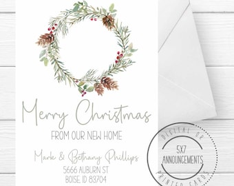 May include: A Christmas card with a watercolour wreath design, featuring pinecones, berries, and greenery. The card reads "Merry Christmas From Our New Home" with a family name and address. A white envelope is included.