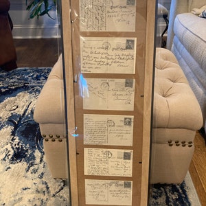 May include: A framed display of vintage postcards with handwritten messages. The postcards are in various shades of cream and tan with black ink. The postcards feature handwritten messages, stamps, and postmarks. The frame is black with a brown mat.