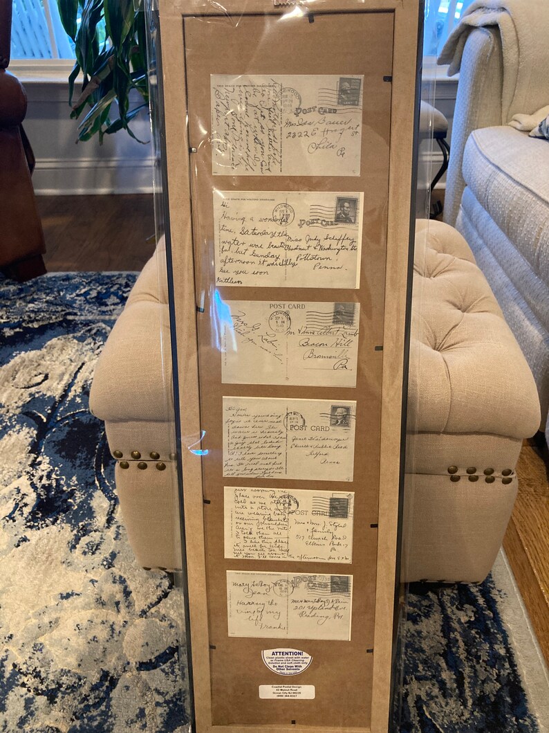 May include: A framed display of vintage postcards with handwritten messages. The postcards are in various shades of cream and tan with black ink. The postcards feature handwritten messages, stamps, and postmarks. The frame is black with a brown mat.