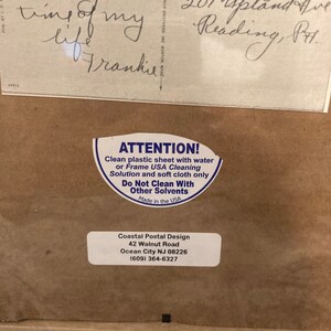 May include: A white sticker with black text that reads "ATTENTION! Clean plastic sheet with water or Frame USA Cleaning Solution and soft cloth only. Do Not Clean With Other Solvents. Made in the USA." Below the sticker is a brown paper background with white text that reads "Coastal Postal Design 42 Walnut Road Ocean City NJ 08226 (609) 364-6327"