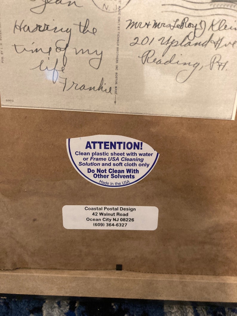 May include: A white sticker with black text that reads "ATTENTION! Clean plastic sheet with water or Frame USA Cleaning Solution and soft cloth only. Do Not Clean With Other Solvents. Made in the USA." Below the sticker is a brown paper background with white text that reads "Coastal Postal Design 42 Walnut Road Ocean City NJ 08226 (609) 364-6327"