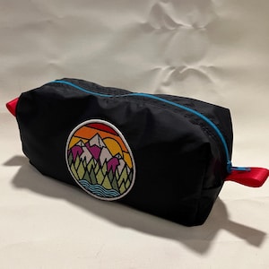 May include: Black zippered travel bag with a colorful mountain scene embroidered on the front. The bag has a red zipper pull and a blue zipper.