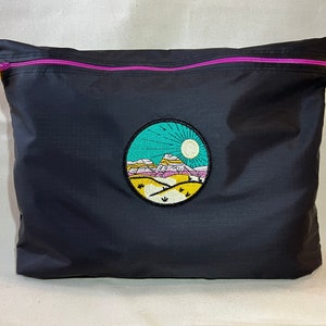 Könnte beinhalten: Eine schwarze Reißverschlusstasche mit einem bunten gestickten Design einer Berglandschaft mit Sonne und türkisfarbenem Himmel. Der Reißverschluss ist pink und die Zuglaschen sind gelb.