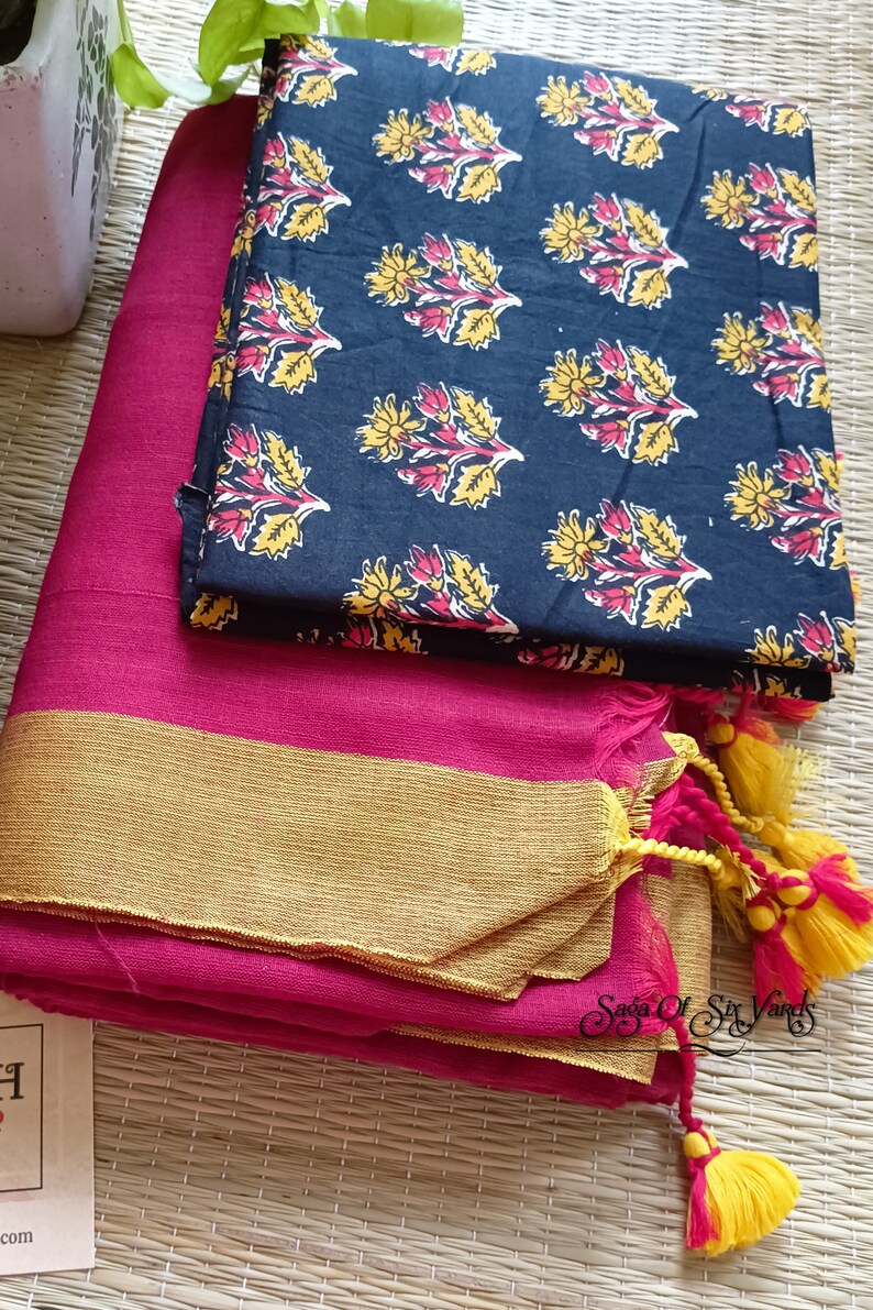 May include: A vibrant pink fabric with a gold border is folded neatly, topped with a navy blue patterned fabric. The navy fabric features a repeating floral design in yellow and red. Yellow and pink tassels are visible.