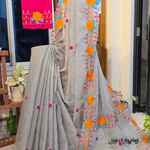 May include: A grey and white chequered saree featuring a vibrant border with orange floral appliques, bright blue accents, and pink embroidered wavy lines. A matching hot pink fabric piece with similar floral embroidery is draped nearby.
