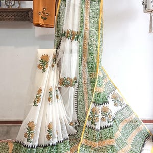 Könnte beinhalten: Weißer Stoff mit einem Blumenmuster in Gelb und Grün. Der Stoff ist über einem Holzständer drapiert. Ein passender orangefarbener Stoff mit floralem Design ist ebenfalls sichtbar.