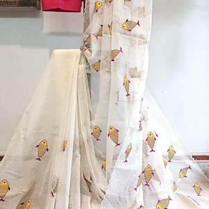 May include: White fabric featuring a repeating pattern of yellow fish with red fins. A matching pink fabric with embroidered fish hangs above. The fabric is draped over a wooden rod. The text "Saga Of Six Yards" is visible.