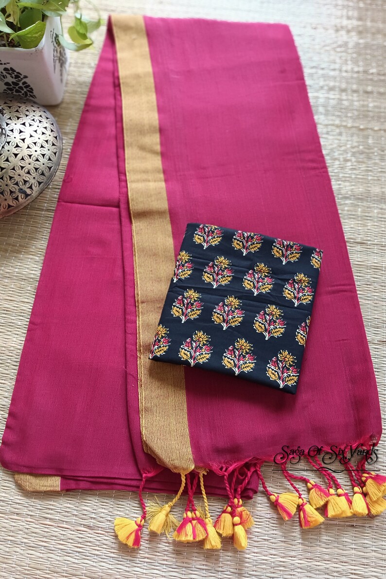 May include: A magenta textile with a gold border and tassels. A black square fabric with a floral pattern in yellow, red, and white rests on top. The text "Saga Of Six Yards" is visible in the bottom right corner.