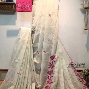 以下が含まれることがあります： 白地に格子柄のピンクの花柄刺繍が施された白い綿のサリー。サリーは波状の縁取りとピンクのブラウスが付いています。