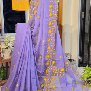 May include: A lavender checkered saree featuring yellow floral embroidery along the scalloped borders and a separate yellow blouse piece with matching lavender floral embroidery, displayed on a wooden stand with the text Saga Of Six Yards.