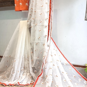 May include: White saree with a delicate floral pattern and a red border. An orange blouse piece with matching embroidery is also included. The saree is draped over a wooden rack, showcasing its intricate design and texture.