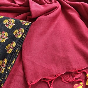 May include: A draped, deep red fabric with a textured appearance, paired with a folded black textile featuring a yellow and red floral pattern. The red fabric has tassels with yellow accents.