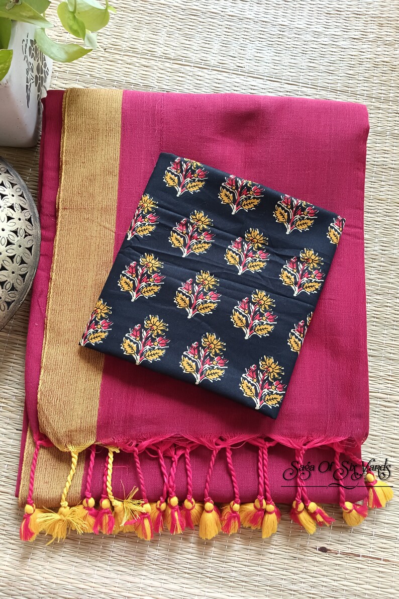 May include: A magenta textile with a gold border and a black patterned fabric. The black fabric has a repeating floral design in yellow and red. The magenta textile has a fringe with yellow and magenta tassels.
