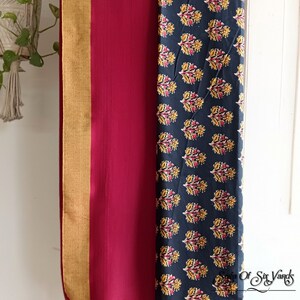 May include: A burgundy and navy blue scarf with a floral pattern and gold trim, hanging on a wooden hanger. The scarf has yellow tassels at the bottom. The right side of the scarf has a navy blue background with a yellow and red floral pattern.