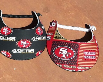 Puede incluir: Dos gorras de visera de los San Francisco 49ers, negras y rojas. Una gorra tiene un fondo negro con logotipos de los 49ers rojos y dorados. La otra gorra tiene un fondo rojo con logotipos de los 49ers dorados y negros y pedrería.