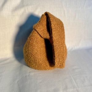 May include: A textured, golden-brown fabric bag with a folded-over top. The bag has a rounded shape and appears to be made of a thick, woven material. The bag is sitting on a white surface.