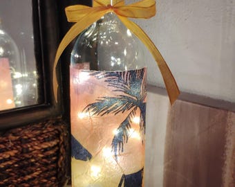 Puede incluir: Una botella de vidrio decorativa con tapón de corcho, envuelta con una escena de playa e iluminada por luces de hadas. La botella está adornada con una cinta amarilla. La escena de la playa presenta una palmera, una silla de playa y una puesta de sol.