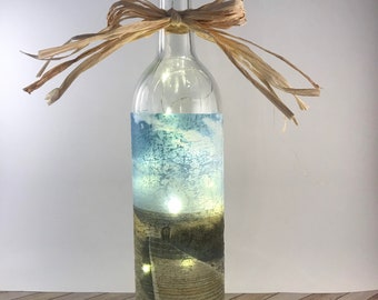 Op de afbeelding: Een heldere glazen fles met een kurken stop en een strik van raffia. De fles is gevuld met witte sprookjeslichtjes en een geprinte afbeelding van een strandscene met een houten loopbrug die naar een vuurtoren leidt.