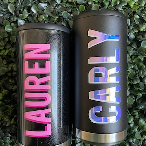 Puede incluir: Dos contenedores de bebidas aislados negros con nombres personalizados. El contenedor de la izquierda tiene el nombre "LAUREN" en letras rosas. El contenedor de la derecha tiene el nombre "CARLY" en letras iridiscentes.