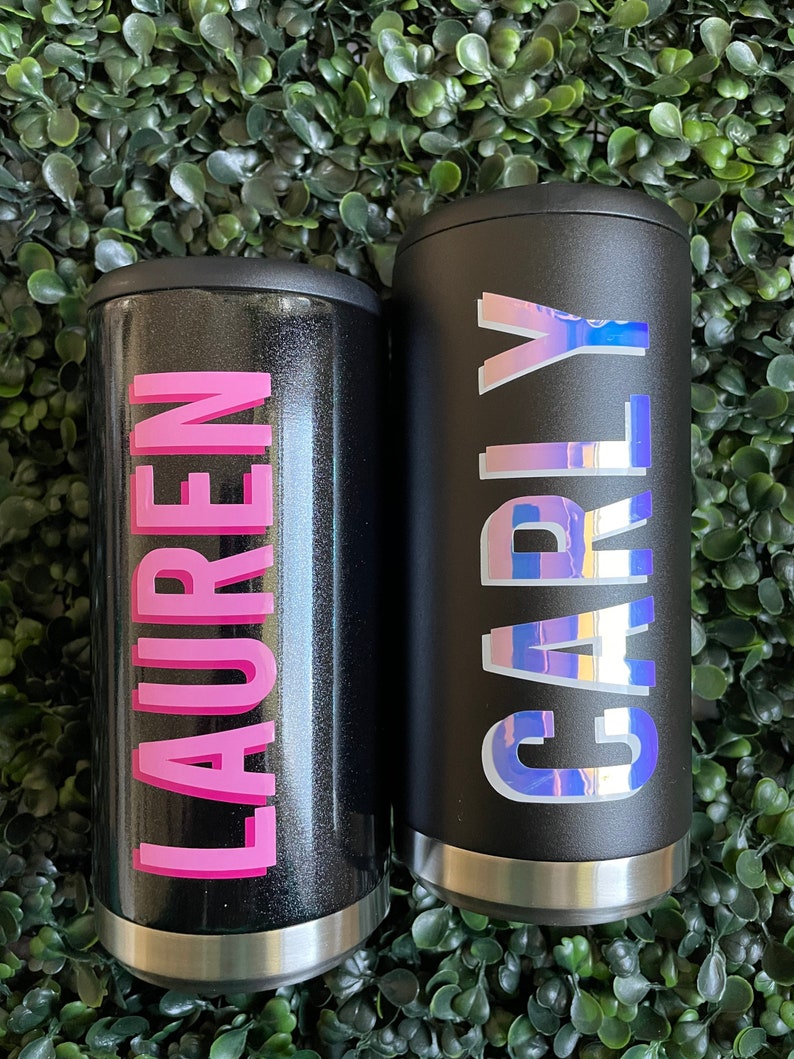 Puede incluir: Dos contenedores de bebidas aislados negros con nombres personalizados. El contenedor de la izquierda tiene el nombre "LAUREN" en letras rosas. El contenedor de la derecha tiene el nombre "CARLY" en letras iridiscentes.