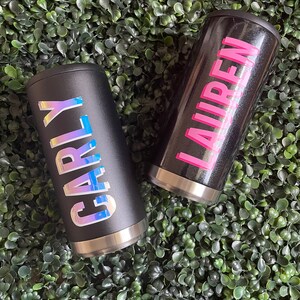 Puede incluir: Dos vasos de acero inoxidable negro con nombres personalizados. El vaso de la izquierda tiene el nombre "Carly" en letras iridiscentes. El vaso de la derecha tiene el nombre "Lauren" en letras rosas.