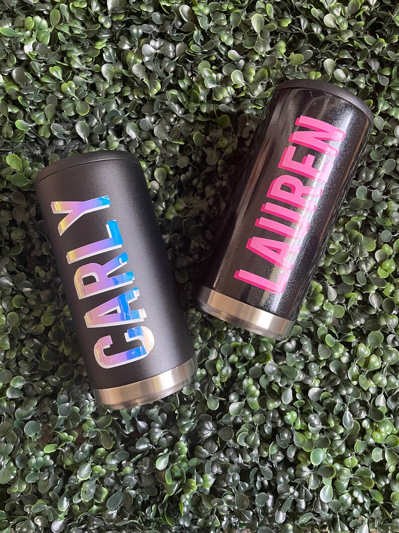 Puede incluir: Dos vasos de acero inoxidable negro con nombres personalizados. El vaso de la izquierda tiene el nombre "Carly" en letras iridiscentes. El vaso de la derecha tiene el nombre "Lauren" en letras rosas.