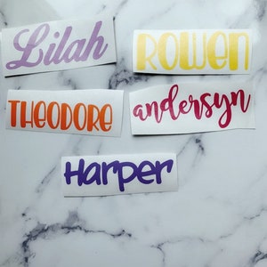 May include: Five personalized name decals in various colors. The names are Lilah in purple, Rowen in yellow, Theodore in orange, andersen in pink, and Harper in purple.