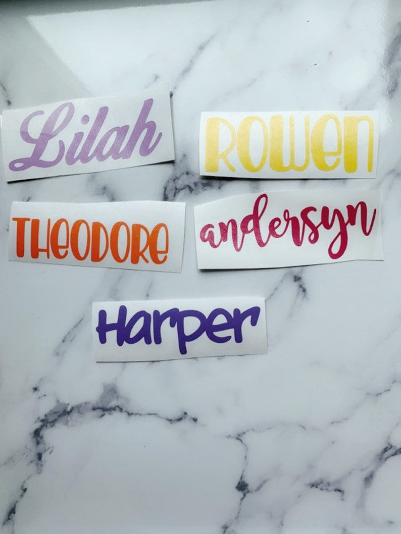 May include: Five personalized name decals in various colors. The names are Lilah in purple, Rowen in yellow, Theodore in orange, andersen in pink, and Harper in purple.