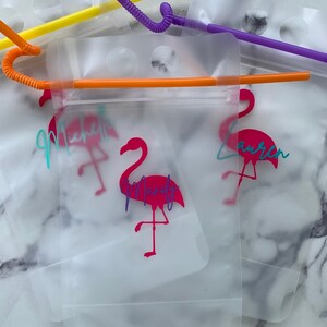 K&ouml;nnte beinhalten: Transparente Trinkbeutel aus Kunststoff mit bunten Strohhalmen. Jeder Beutel hat ein rosa Flamingo-Design und einen Namen in Schreibschrift. Die Strohhalme sind gelb, orange und lila. Die Beutel sind f&uuml;r Getr&auml;nke bestimmt.