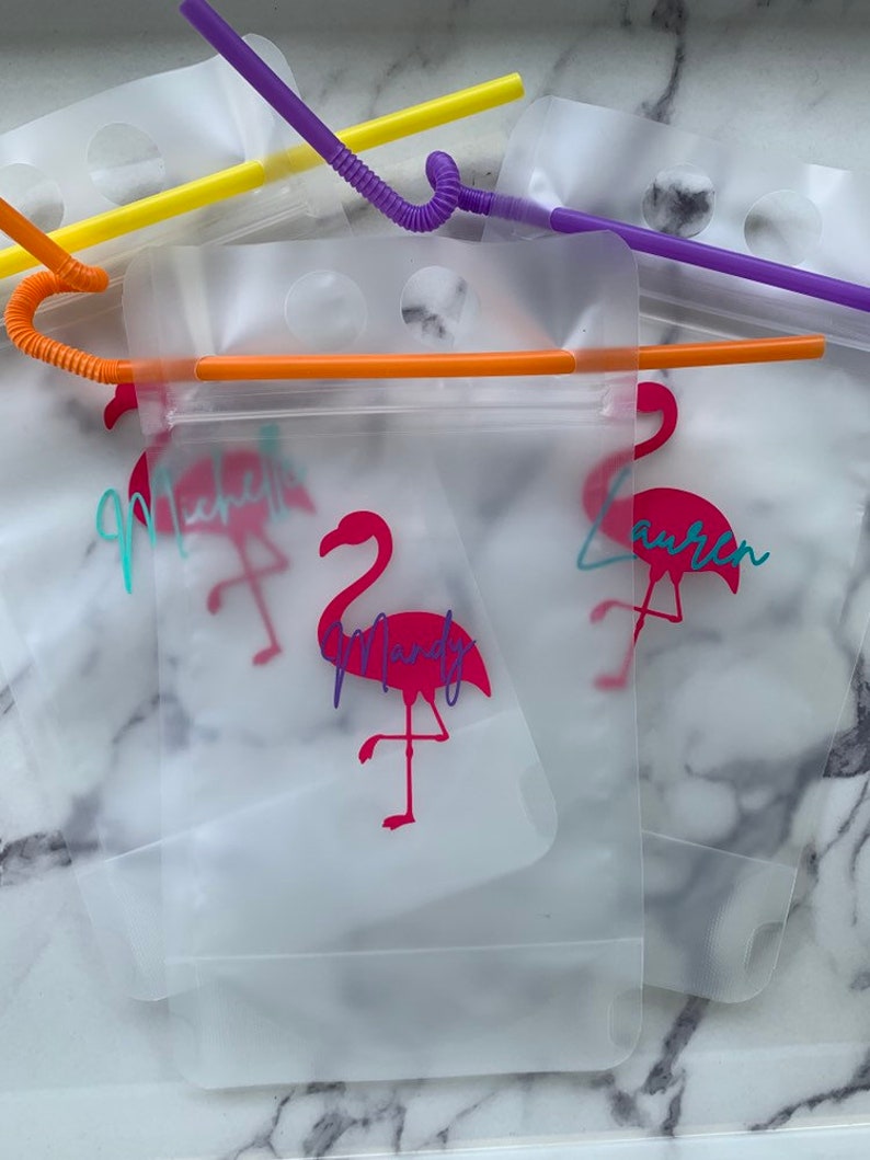 K&ouml;nnte beinhalten: Transparente Trinkbeutel aus Kunststoff mit bunten Strohhalmen. Jeder Beutel hat ein rosa Flamingo-Design und einen Namen in Schreibschrift. Die Strohhalme sind gelb, orange und lila. Die Beutel sind f&uuml;r Getr&auml;nke bestimmt.