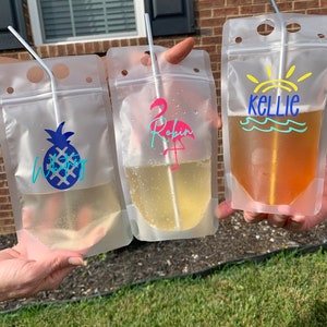 K&ouml;nnte beinhalten: Drei transparente Getr&auml;nkebeutel mit Strohhalmen, die jeweils ein anderes Getr&auml;nk enthalten. Einer hat ein blaues Ananas-Design, ein anderer einen rosa Flamingo und den Namen "Robin", der dritte den Namen "Kellie" mit einem Sonnen- und Wellen-Design.