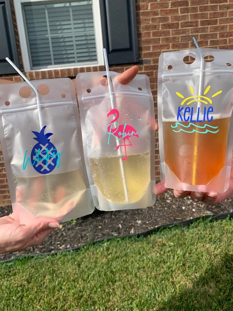 K&ouml;nnte beinhalten: Drei transparente Getr&auml;nkebeutel mit Strohhalmen, die jeweils ein anderes Getr&auml;nk enthalten. Einer hat ein blaues Ananas-Design, ein anderer einen rosa Flamingo und den Namen "Robin", der dritte den Namen "Kellie" mit einem Sonnen- und Wellen-Design.