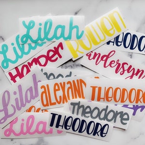 May include: A collection of colorful vinyl stickers with various names written in a cursive font. The names include Lilah, Harper, Ruen, Anderson, Theodore, and Alexand.