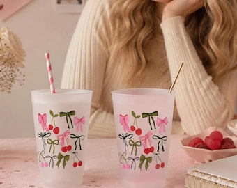 May include: Two frosted plastic tumblers featuring a repeating pattern of small red cherries and bows in shades of pink, green, and black. One tumbler contains a pink and white striped straw, while the other holds a thin gold-colored metal straw.