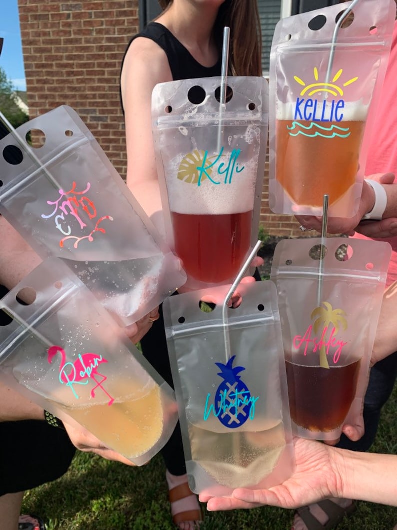 K&ouml;nnte beinhalten: Transparente Getr&auml;nkebeutel aus Kunststoff mit verschiedenen bunten Designs und Namen, darunter "Robin", "Ashley", "Whitney" und "Kellie". Jeder Beutel enth&auml;lt ein andersfarbiges Getr&auml;nk und einen Edelstahlstrohhalm. Die Beutel werden von mehreren Personen gehalten.