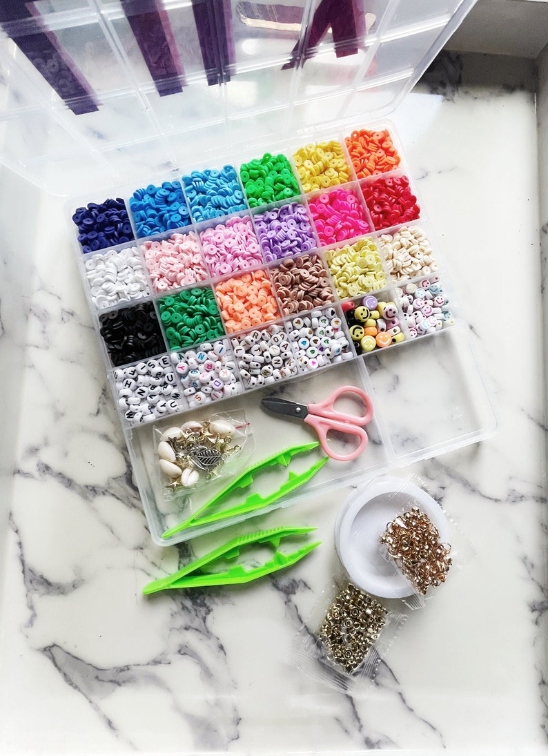 May include: A clear plastic organiser filled with colourful beads, including letter beads and smiley face beads. Also included are pink scissors, green tweezers, and small bags of jewellery-making supplies. The beads are arranged in a variety of colours.