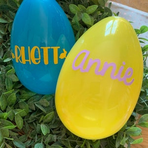 May include: Two plastic Easter eggs, one blue with the name "Rhett" in yellow letters and one yellow with the name "Annie" in purple letters.