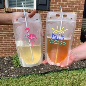 K&ouml;nnte beinhalten: Zwei transparente Plastikbeutel, gef&uuml;llt mit gelber und bernsteinfarbener Fl&uuml;ssigkeit, jeweils mit einem Strohhalm. Ein Beutel hat einen rosa Flamingo und den Namen "Robin" aufgedruckt. Der andere Beutel hat eine Sonne und den Namen "Kellie".