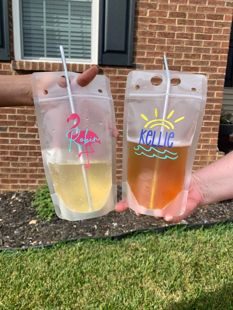 K&ouml;nnte beinhalten: Zwei transparente Plastikbeutel, gef&uuml;llt mit gelber und bernsteinfarbener Fl&uuml;ssigkeit, jeweils mit einem Strohhalm. Ein Beutel hat einen rosa Flamingo und den Namen "Robin" aufgedruckt. Der andere Beutel hat eine Sonne und den Namen "Kellie".