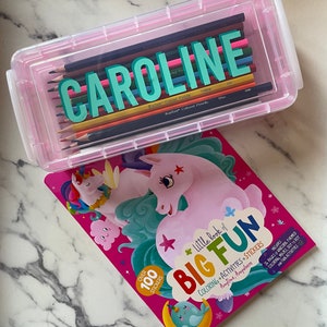 May include: A pink plastic pencil case with the name "CAROLINE" written on it. The case is filled with colored pencils. A pink and blue unicorn coloring book with the title "Little Book of Big Fun" is next to the case.
