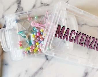 May include: A clear plastic craft box with the name "Mackenzie" in purple and white lettering. The box is open, revealing colorful beads, charms, and string. The beads are in various pastel colors, including pink, yellow, and blue.